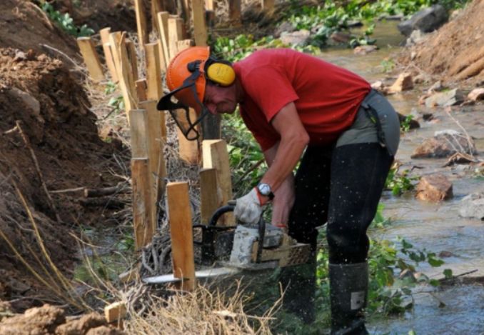 Un ouvrier du génie écologique, équipé d'un casque de protection et des gants effectue des travaux de renforcement de la berge d'une rivière